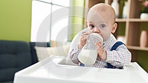 Adorable caucasian baby drinking feeding bottle sitting on highchair at home