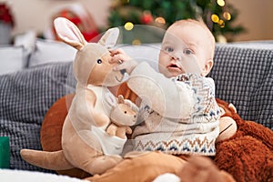 Adorable caucasian baby holding kangaroo doll sitting on sofa by christmas tree at home