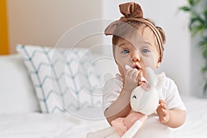 Adorable caucasian baby sitting on bed bitting doll at bedroom