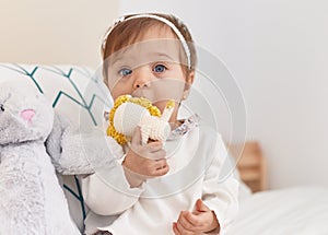 Adorable caucasian baby sitting on bed bitting doll at bedroom