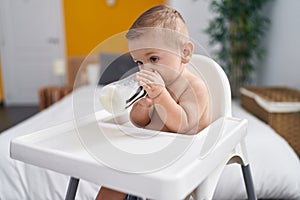 Adorable caucasian baby sucking milk on feeding bottle sitting on highchair at home