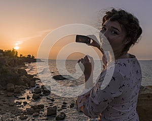 Adorable Caucasian Brunette girl in Light Dress Enjoying with Phone at the Seacoast at Sunrise time.