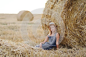 adorable caucasian girl enjoy having fun sitting near golden hay bale on wheat harvested field near farm.