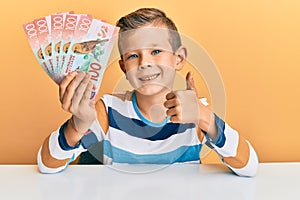 Adorable caucasian kid holding 100 new zealand dollars banknote sitting on the table smiling happy and positive, thumb up doing