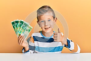Adorable caucasian kid holding israel 50 shekels sitting on the table smiling happy and positive, thumb up doing excellent and