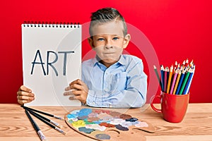 Adorable caucasian kid painter sitting at art studio holding art word skeptic and nervous, frowning upset because of problem
