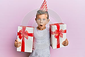 Adorable caucasian kid wearing birthday hat holding presents afraid and shocked with surprise and amazed expression, fear and