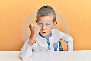 Adorable caucasian kid wearing casual clothes sitting on the table angry and mad raising fist frustrated and furious while