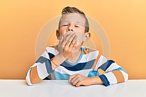 Adorable caucasian kid wearing casual clothes sitting on the table bored yawning tired covering mouth with hand