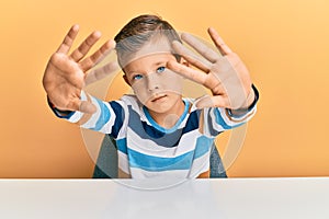 Adorable caucasian kid wearing casual clothes sitting on the table doing frame using hands palms and fingers, camera perspective
