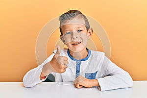 Adorable caucasian kid wearing casual clothes sitting on the table doing happy thumbs up gesture with hand
