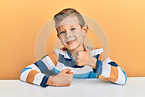 Adorable caucasian kid wearing casual clothes sitting on the table doing happy thumbs up gesture with hand