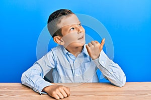 Adorable caucasian kid wearing casual clothes sitting on the table pointing thumb up to the side smiling happy with open mouth