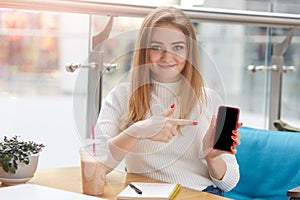 Adorable cheerful young Caucasian woman holds smart phone and points fore finger at black display, showing copyspace  over