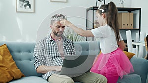 Adorable child decorating father with bijouterie while man is working with laptop at home