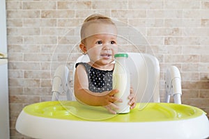 Adorable child drinking from bottle. funny baby eating healthy food on kitchen. Baby girl sitting in high chair and holding a