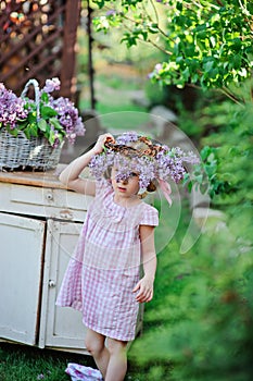 Adorable child girl wearing lilac wreath in pink plaid dress near vintage bureau in spring garden