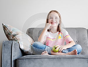 Adorable child happy girl 8 years old sits on a gray sofa with a green apple in hands