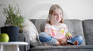 Adorable child girl 8 years old sits on a gray sofa with a green apple and a TV remote control in her hands rest and entertainment