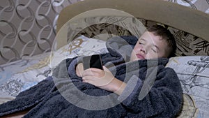 Adorable Child in Home Gray Dressing Gown Falls Asleep with Smartphone in Hands