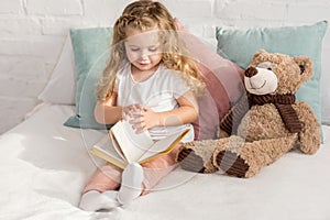 adorable child playing with teddy bear and reading book on bed