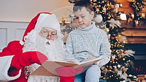 Adorable child and Santa Claus reading book near Christmas tree and talking on New Year& x27;s day