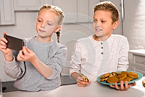 adorable child taking selfie on smartphone with brother holding plate with delicious cookies