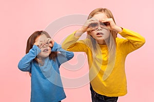 Adorable, curious, nosy two little girls looking through fingers imitating binoculars, exploring world