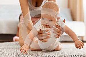 Adorable cute baby in white bodysuit crawling on floor on carpet while mother is helping and supporting, posing in light room at