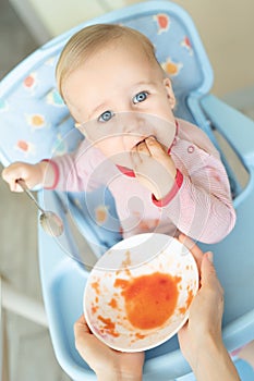 Adorable cute caucasian blond toddler boy eating tasty vegetable soup in chair at kithcen indoor. Mother feeding baby healthy food