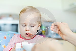 Adorable cute caucasian blond toddler boy eating tasty vegetable soup in chair at kithcen indoor. Mother feeding baby healthy food