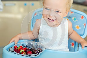 Adorable cute caucasian blond toddler boy enjoy tasting different seasonal fresh ripe organic berries sitting in highchair at home