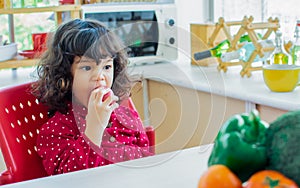 Adorable cute Caucasian little girl wearing casual clothes, eating and biting red apple for breakfast in kitchen at home with