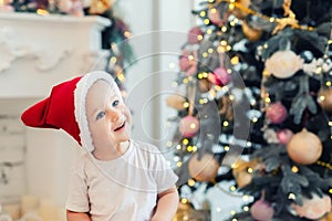 Adorable cute child boy having fun playing at home in red santa claus hat and festive christmas tree on background. Happy toddler