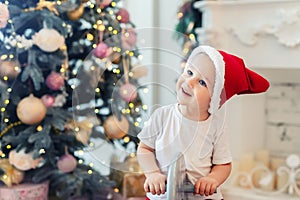 Adorable cute child boy having fun plying at home in red santa claus hat and festive christmas tree on background. Happy