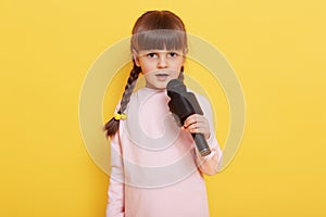 Adorable cute child with microphone in hands singing songs, looks at camera, performing isolated over yellow background, child