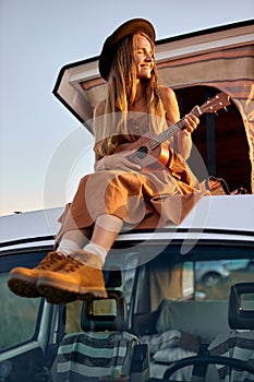 Adorable cute lady in dress and hat sitting on top of minig retro van playing ukulele