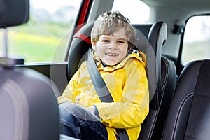 Adorable cute preschool kid boy sitting in car in yellow rain coat. Little school child in safety car seat with belt