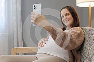 Adorable delighted brown haired pregnant woman smiling while taking selfie with her phone expectant mother making photo capturing