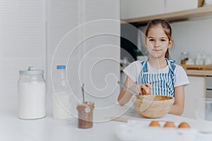 Adorable European child enjoys cooking activity, breaks egg in bowl, whisks ingredients, prepares tasty pastry, being young cook,