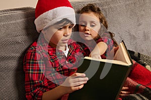 Adorable European kids, brother in Santa`s hat and his younger sister sitting on the couch and reading a book, fairy tales on the