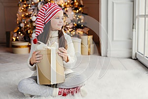 Adorable female child wears santa claus hat, warm clothes, hold