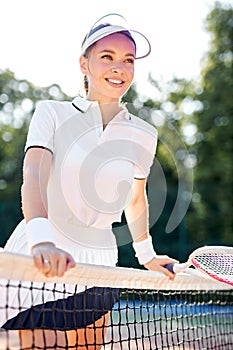 Adorable female tennis player in white uniform stands next to net posing with racket