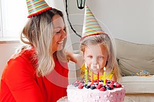 Adorable four years old european blonde girl makes a wish before blowing out the candles on a birthday cake that mom is holding