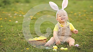 Adorable funny baby boy with bunny ears playing on green grass with Easter eggs. The child laughs merrily. Easter