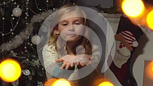 Adorable girl is blowing artificial snow on her hands against a Christmas tree