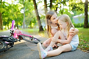 Adorable girl comforting her little sister after she fell off her bike at summer park. Child getting hurt while riding a bicycle.