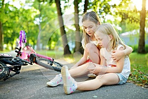 Adorable girl comforting her little sister after she fell off her bike at summer park. Child getting hurt while riding a bicycle.