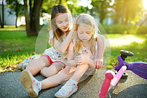 Adorable girl comforting her little sister after she fell off her scooter at summer park. Child getting hurt while riding a kick