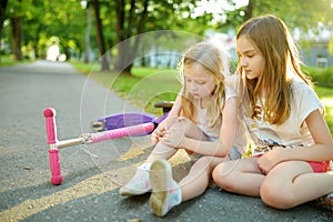 Adorable girl comforting her little sister after she fell off her scooter at summer park. Child getting hurt while riding a kick s